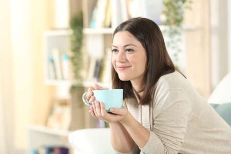 Satisfied woman thinking and drinking coffee looking away sitting on a sofa at homeの写真素材