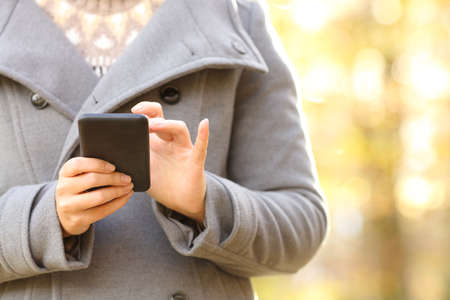 Close up front view of woman hands using a smart phone in autumn or winter in a parkの写真素材
