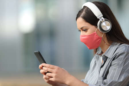 Side view portrait of a happy woman wearing headphones and mask listening to music from smart phoneの写真素材