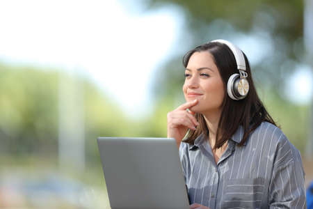 Satisfied woman with laptop and wireless headphones thinking looking at side in a parkの写真素材
