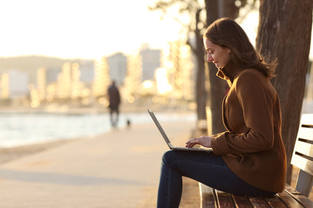 Profile of woman using laptop sitting on a bench in winter in a park in a beach townの写真素材