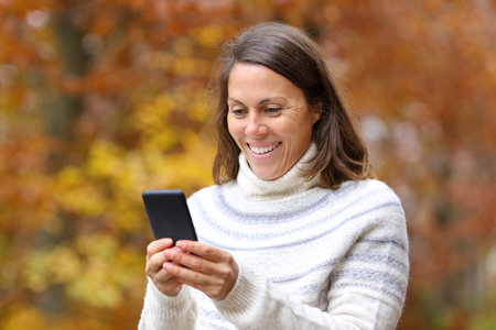 Happy middle age woman using smart phone walking in fall season in a parkの写真素材