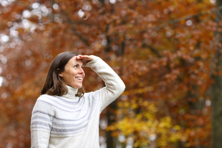 Happy adult woman searching with hand on forehead in a forest in autumnの写真素材