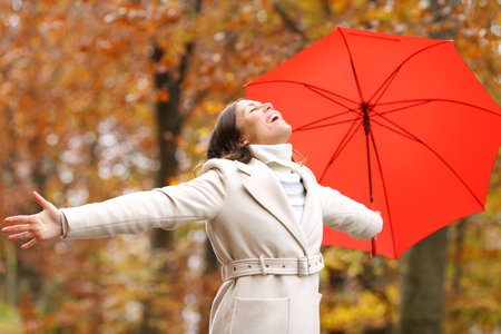 Happy fashion adult woman with umbrella celebrating autumn stretching arms in a parkの写真素材