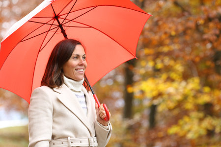 Happy adult woman contemplating autumn under red umbrella in a parkの写真素材