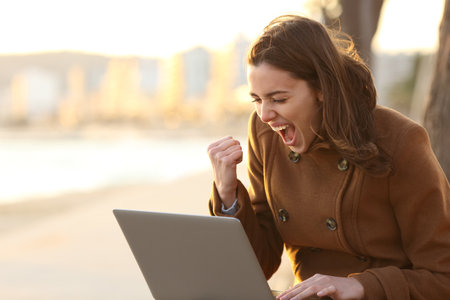 Excited woman reading good news on laptop sitting on a bench in winter on the beachの写真素材