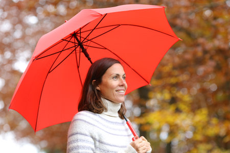 Happy adult woman under red umbrella contemplating views in autumn in a parkの写真素材