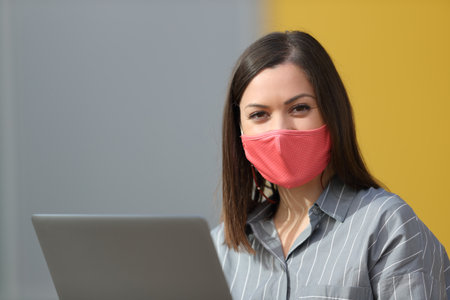 Satisfied woman wearing protective mask and using laptop looking at camera in the streetの写真素材
