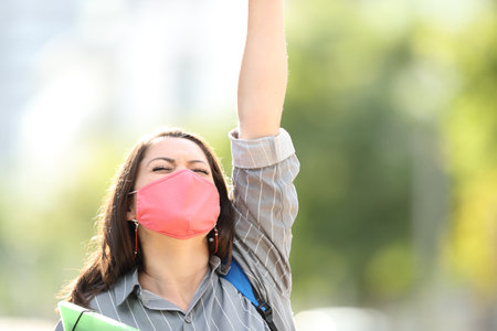 Excited student with mask celebrating success raising arm outdoors in a park