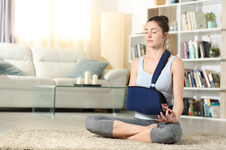 Full body portrait of a disabled woman with a broken arm in a sling practicing yoga exercise at homeの写真素材