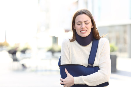 Front view portrait of a stressed woman suffering with broken arm on a sling walking in the streetの写真素材