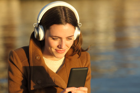 Satisfied woman listening to music with headphones and mobile phone on the beach at sunset in winterの写真素材