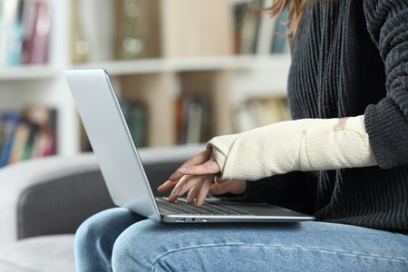 Close up of a disabled woman with bandaged arm using laptop at homeの写真素材