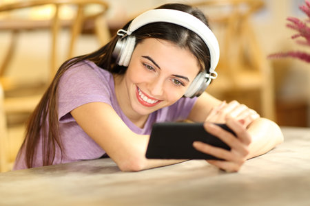 Happy female watching videos on smart phone wearing headphones at homeの写真素材