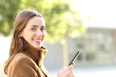 Happy woman holding mobile phone looks at you in winter in the streetの写真素材