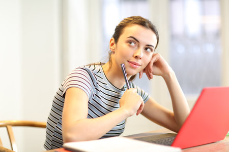 Happy student thinking looking at side with a laptop and notes at homeの写真素材