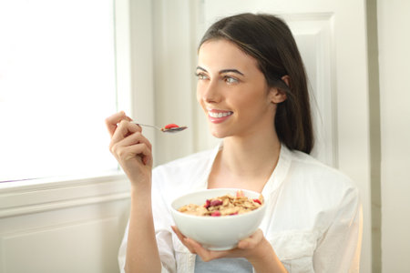 Happy woman eating cereals at breakfast in the morning looking through a window at homeの写真素材