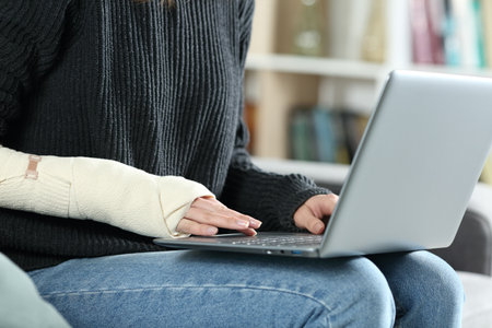 Close up portrait of a disabled woman hands with bandaged arm writing on laptop at homeの写真素材