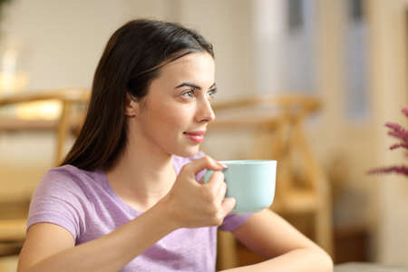 Relaxed woman looking away drinking coffee sitting at homeの写真素材