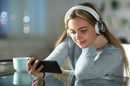 Teen watching videos on smart phone wearing headphones in the night in the living room at homeの写真素材