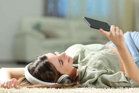 Happy woman listening to music with smart phone and headphones lying on a carpet at homeの写真素材