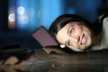 Happy woman late hours in the night checking smart phone content at homeの写真素材