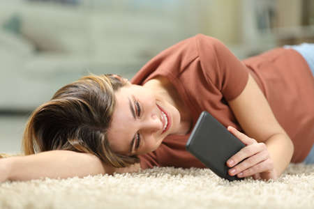 Happy woman reading message on mobile phone lying on a carpet on the floor at homeの写真素材