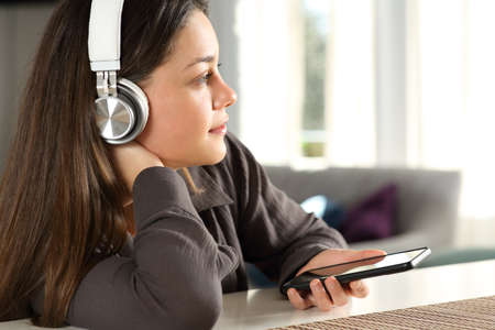 Pensive woman wearing wireless headphones contemplating listening to music at homeの写真素材