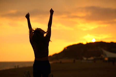 Back view portrait of a woman silhouette raising arms jumping celebrating at sunset on the beachの写真素材