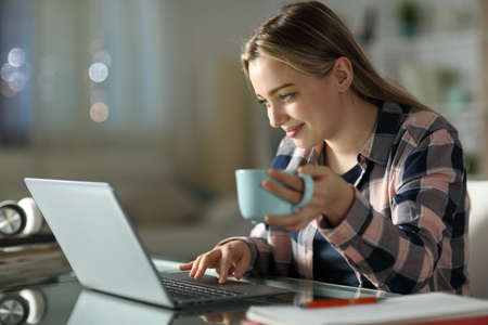 Satisfied student e-learning using laptop holding coffee cup at home in the nightの写真素材