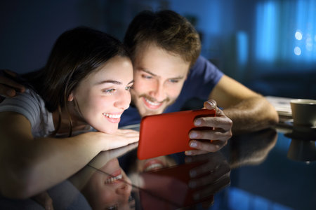 Happy couple watching media content on horizontal mobile phone in the night at homeの写真素材