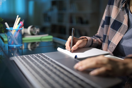 Close up of a student hand taking notes checking laptop in the night at homeの写真素材