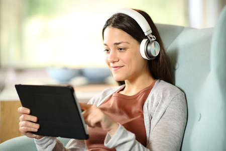 Satisfied woman wearing wireless headphones watching media on tablet on a couch at homeの写真素材