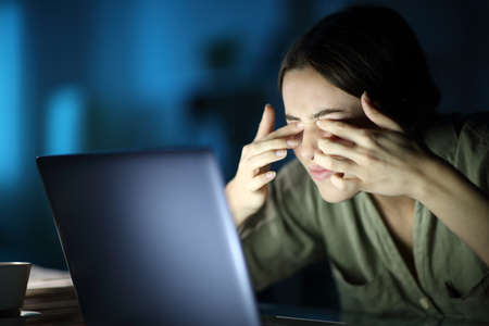 Fatigued woman with eyestrain scratching eyes using laptop late hours in the night at homeの写真素材