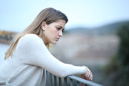 Sad woman looking down in a house balcony complaining alone in a townの写真素材