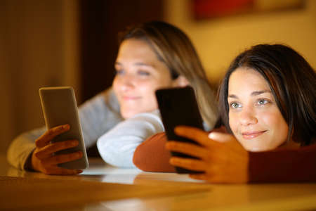 Two satisfied roommates ignoring each other using everyone her smart phone in the night at homeの写真素材