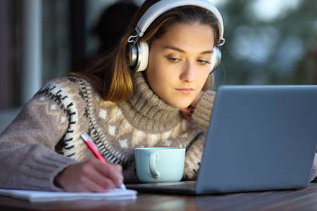 Concentrated student wearing headphones e-learning with laptop taking notes on notebook in a coffee shop in winterの写真素材