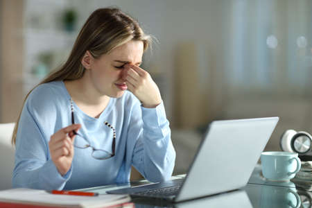 Student suffering eyestrain holding eyeglasses learning with a laptop late hours in the night at homeの写真素材