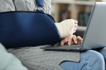 Close up of a woman with broken arm wearing sling using laptop sitting at homeの写真素材