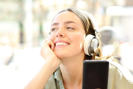 Happy woman wearing headphones and holding a smart phone listening to music dreaming in a coffee...