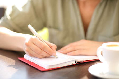 Front view close up of a woman hand writing on paper agenda in a coffee shopの写真素材
