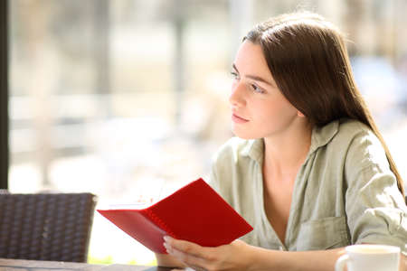 Pensive woman wondering looking away holding a paper agenda in a coffee shopの写真素材