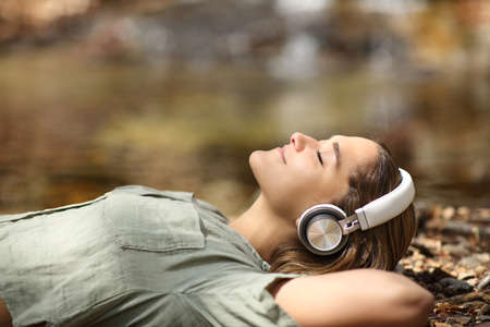 Side view portrait of a relaxed woman wearing headphones listening to music lying in a riversideの写真素材