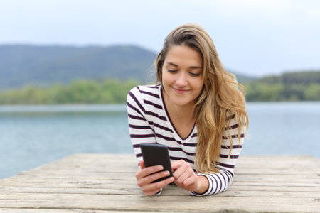 Front view of a happy woman using smartphone lying in a lake pierの写真素材