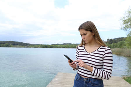 Sad woman reading message on smart phone on vacation in a lakeの写真素材