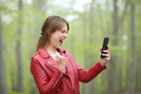 Amazed woman celebrating success checking smart phone in a green parkの写真素材