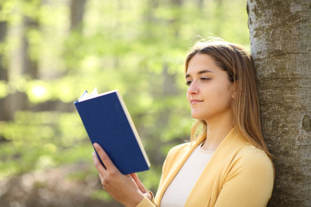 Relaxed woman reading a paper book leaning on a tree in a beautiful forestの写真素材