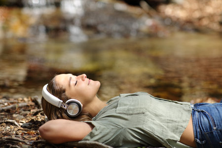 Side view portrait of a happy woman resting lying listening to music wearing wireless headphones in a riversideの写真素材