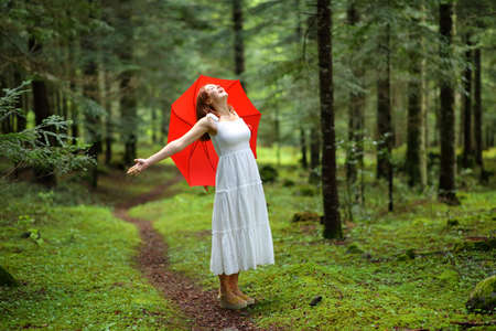Excited woman with red umbrella celebrating vacation in a forest under the rainの写真素材