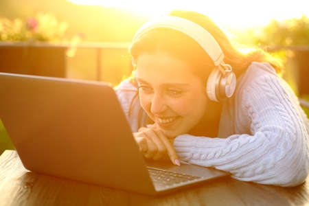 Happy lady with wireless headphones at sunset checking laptop in a balconyの写真素材